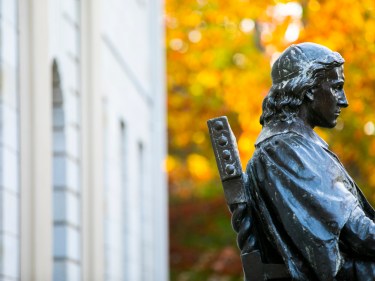 John Harvard statue in front of yellow fall leaves.