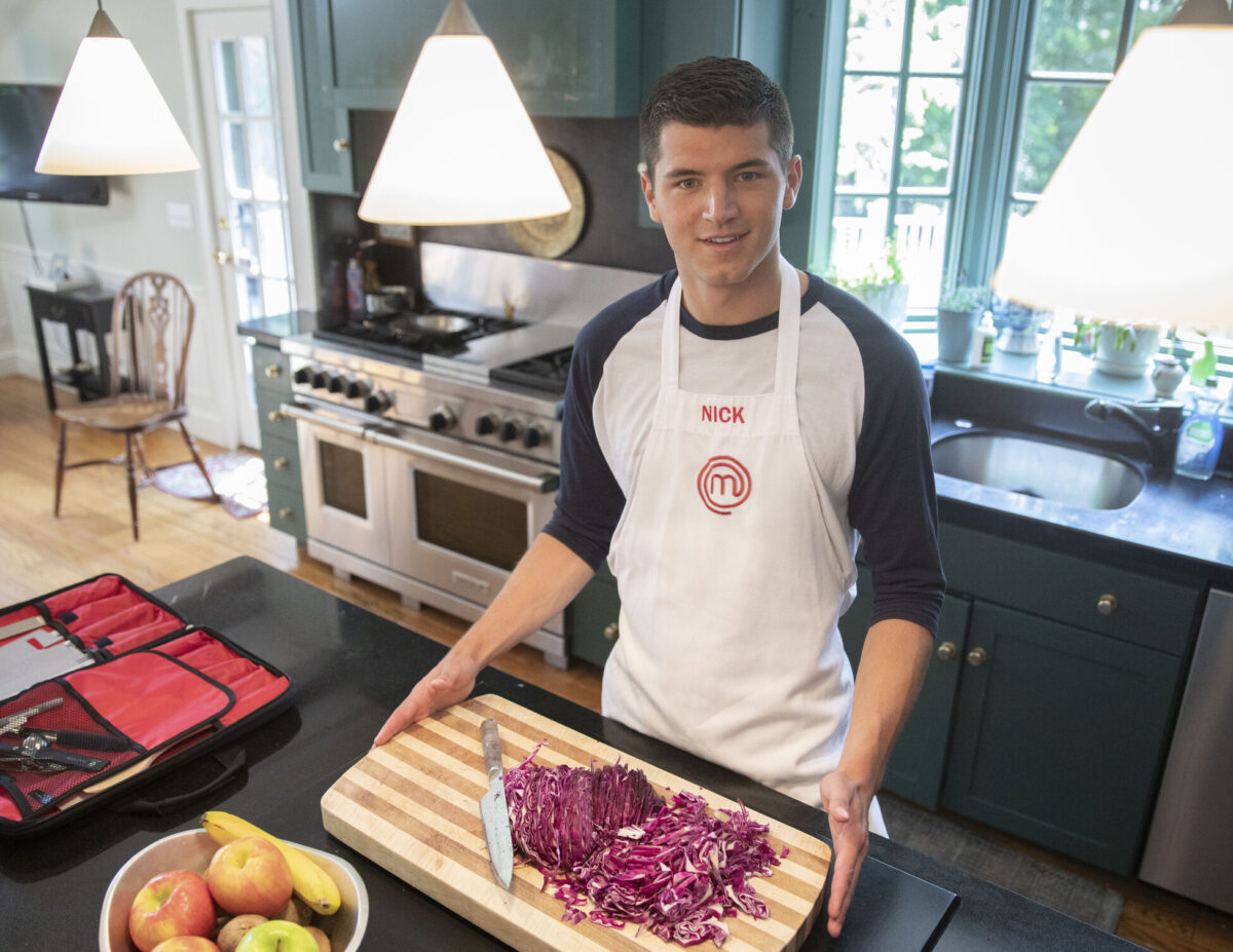A student wearing an apron stands in a kitchen with a cutting board of purple cabbage in front of him