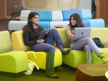 Two students sit on couches and chat while doing school work