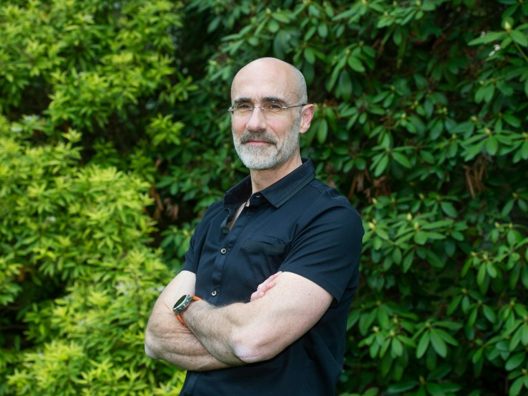 A man stands with his arms crossed in front of greenery